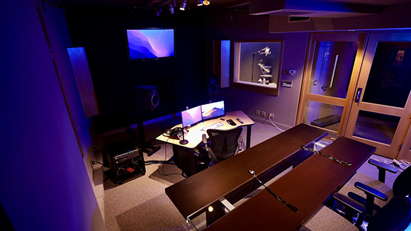 A recording studio control room with a mixing desk, monitors, speakers, and blue ambient lighting.