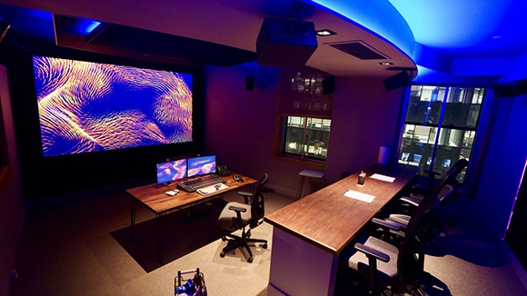 A dark control room featuring a large screen displaying colorful patterns, two desks, and computer monitors.