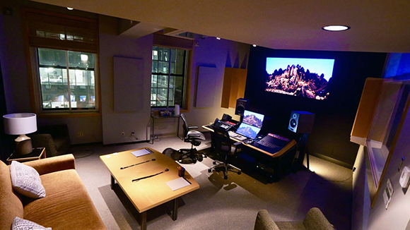A dark editing suite featuring a large projection screen, computer monitors, and a lounge area.