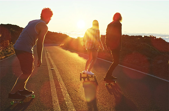 Three people longboarding down a paved road toward a bright sunset over a landscape.