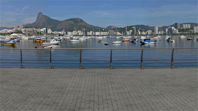 Many small boats float in a calm bay in front of a city skyline and large mountains under a blue sky.