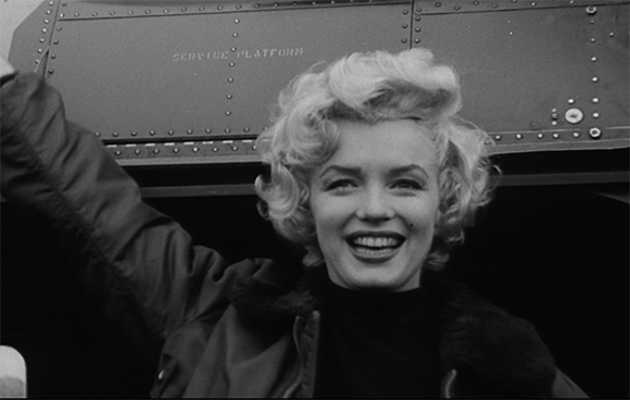 A smiling blonde woman with wavy hair wearing a dark jacket in front of a metal service platform.