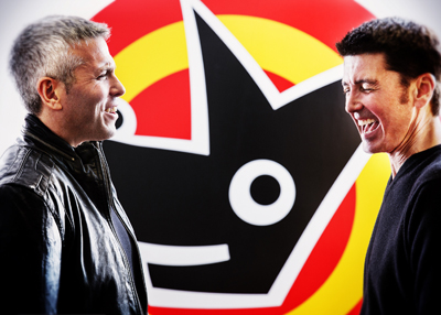 Two men smiling and looking at each other in front of a large red, yellow, and black graphic logo.