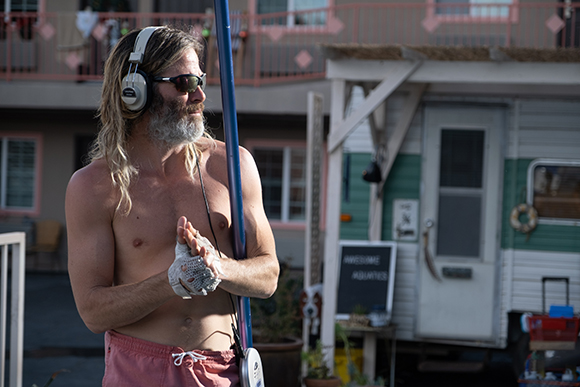 Shirtless man with long blonde hair, sunglasses, and headphones stands near a mobile home.