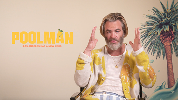 A man with a grey beard and blonde hair wears a yellow cardigan and gestures with both hands.