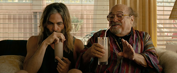 A man with long hair and a man with glasses and a goatee sit on a couch drinking milkshakes.