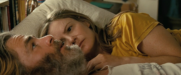 A man with a grey beard and a woman in a yellow shirt lie close together on pillows.