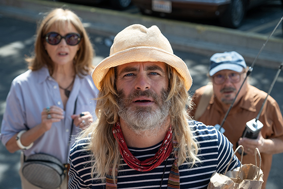 A man with long blonde hair and a bucket hat looks forward, flanked by a woman and a man.