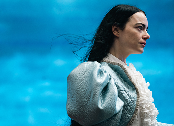 A woman with long dark hair wearing a light blue ruffled jacket against a blue background.