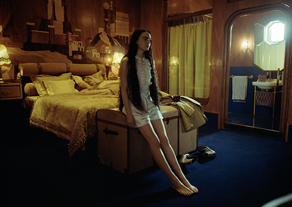 A woman with long dark hair sits on a trunk in a dimly lit, vintage-style bedroom.