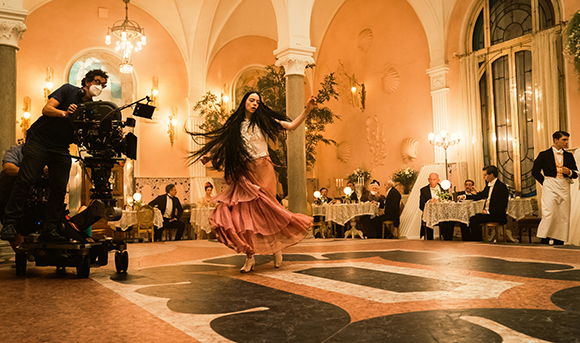A woman in a long pink skirt dances in a large, warm-lit ballroom while a camera crew films her.