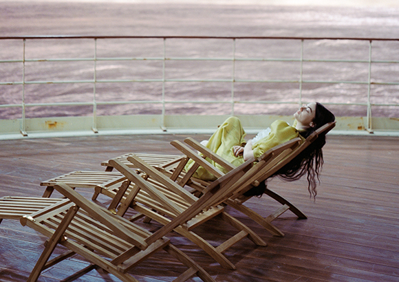 A person in a yellow outfit reclines in a wooden lounge chair on a ship deck overlooking the ocean.