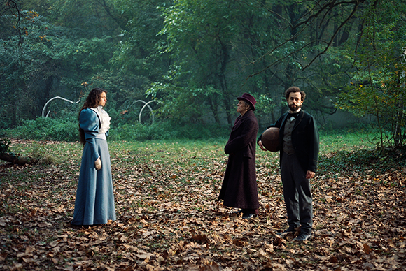 Three people in vintage attire stand in a wooded area filled with fallen autumn leaves.