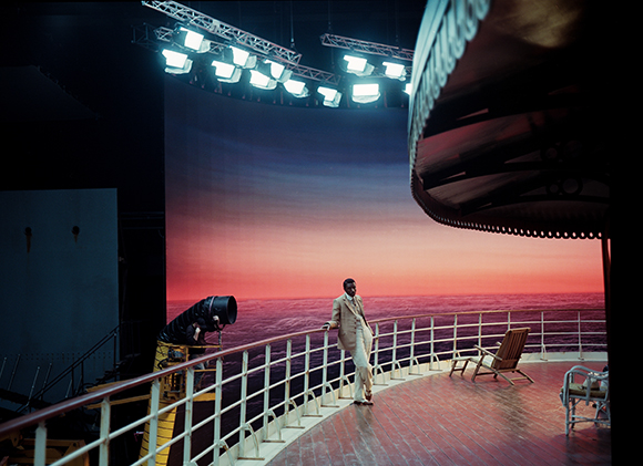 A person in light clothing stands on a ship's deck overlooking a sunset ocean under studio lighting.