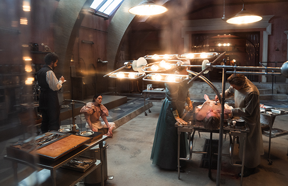 Medical staff in period clothing working in a dark, vaulted stone operating room with bright surgical lights.