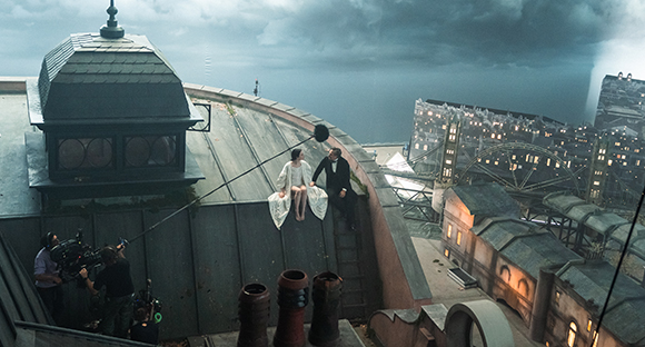 Film crew with a camera filming two actors sitting on a rooftop edge against a dark, cloudy cityscape.