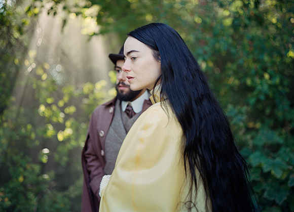 A woman with long black hair in a yellow top stands in a forest with a man behind her.