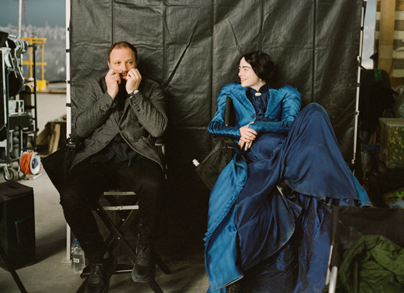A man in a grey suit and a woman in a blue period gown sit side-by-side against a black backdrop.