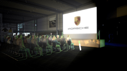 An audience sits in a dark room watching a large bright screen displaying the Porsche logo.