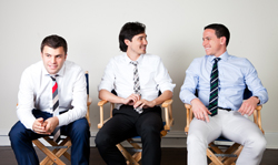 Three men in dress shirts and ties sitting in wooden director's chairs against a white background.