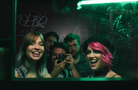 Five young people smiling and posing behind a glass pane under dim green lighting.