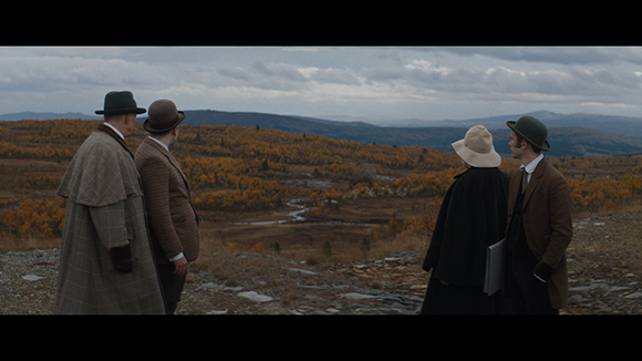 Four people in vintage coats and hats look out over a valley of autumn trees under a cloudy sky.