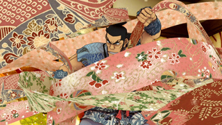 An animated man with dark hair and blue armor surrounded by swirling, colorful patterned fabric strips.