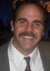 A smiling man with dark hair and a mustache wearing a grey suit, white shirt, and patterned tie.