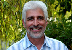 A smiling man with short gray hair and a gray beard wearing a light blue striped shirt outdoors.