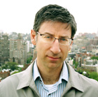 A man with short dark hair and glasses wearing a light jacket and striped shirt against a city backdrop.