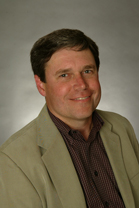 A smiling man with brown hair wearing a tan blazer over a dark patterned button-down shirt.