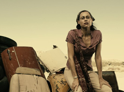 A woman in a purple shirt and glasses sits on junk in a barren, yellowish landscape.