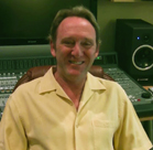 A smiling man in a yellow shirt sitting in front of a mixing console and studio monitors.