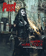 Woman in dark, hooded costume with metallic accents stands in a workshop with sparks flying from machinery.
