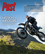 A person on a dirt bike jumps in the air with mountains and a blue sky in the background.