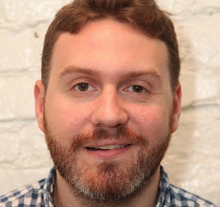 A man with reddish-brown hair and a beard smiling slightly against a white brick wall.