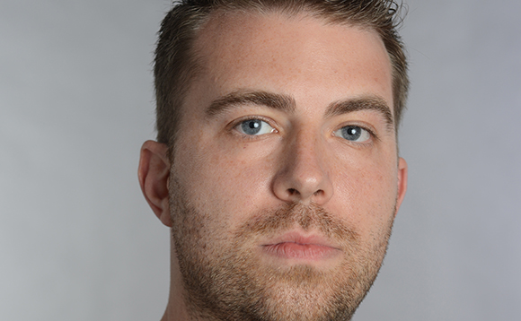 Close-up portrait of a man with light blue eyes and short brown hair against a gray background.