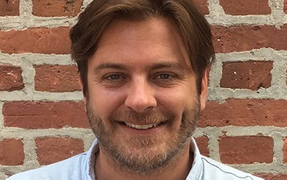 A smiling man with brown hair and a beard standing in front of a red brick wall.