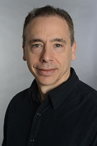 A man with short brown hair and a black button-down shirt looking forward against a gray background.