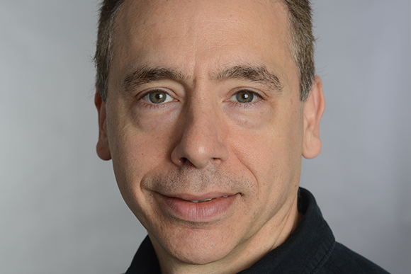 Close-up portrait of a man with short brown hair and green eyes looking forward.