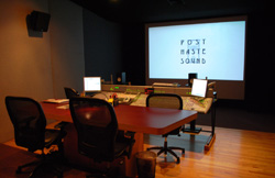 A dark sound studio featuring a large mixing console, computer monitors, and a projection screen.