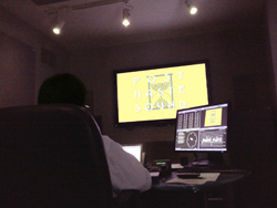 A person sits in a dark room facing a large screen displaying yellow text and a smaller monitor with audio software.