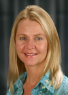 A smiling woman with long blonde hair wearing a blue patterned shirt against a dark background.