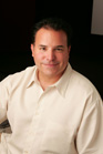 A man with dark hair wearing a cream-colored button-down shirt smiling against a dark background.
