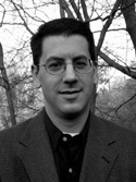 Black and white portrait of a man with short dark hair and glasses wearing a blazer.