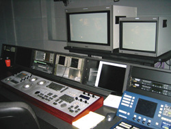 A control desk with multiple monitors, a large mixing console, and various electronic broadcasting equipment.