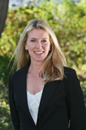 A blonde woman in a black blazer and white top smiling against a background of green foliage.