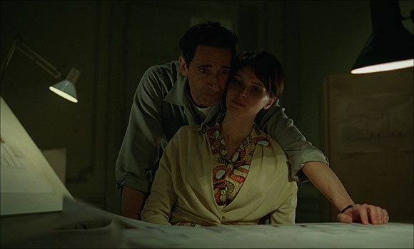 A man leans over a woman, embracing her from behind in a dark room with desk lamps.