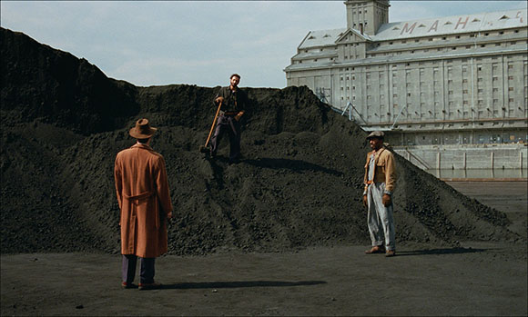 Three men stand near a large mound of dark coal or dirt in front of a large industrial building.