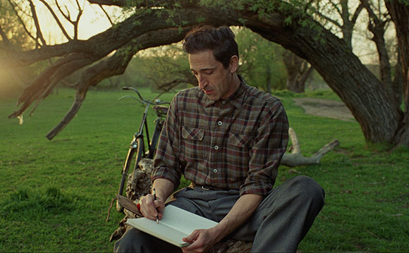 A man in a plaid shirt sits on a log in a grassy field, writing in a notebook under a large tree.
